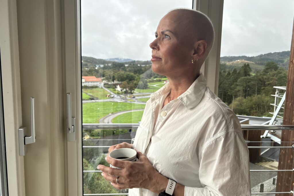 En person med barbert hode og hvit skjorte står ved et vindu med et krus i hånden og ser ut på et naturskjønt, grønt landskap.