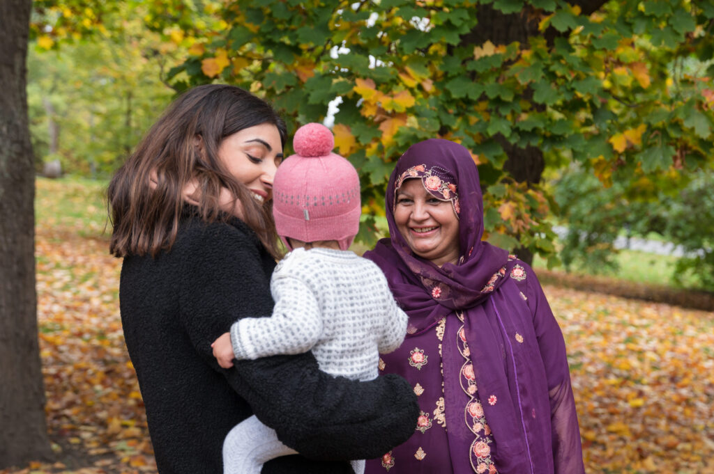 Tre personer, deriblant en kvinne med en baby i armene og en annen kvinne med lilla skaut, står ute i en park med høstløv og trær i bakgrunnen.