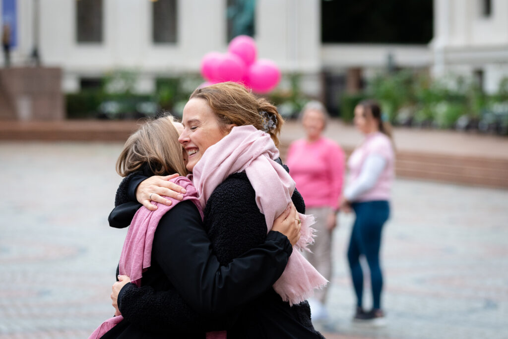 To kvinner med skjerf om halsen omfavner hverandre utendørs, med rosa ballonger og to personer i bakgrunnen.