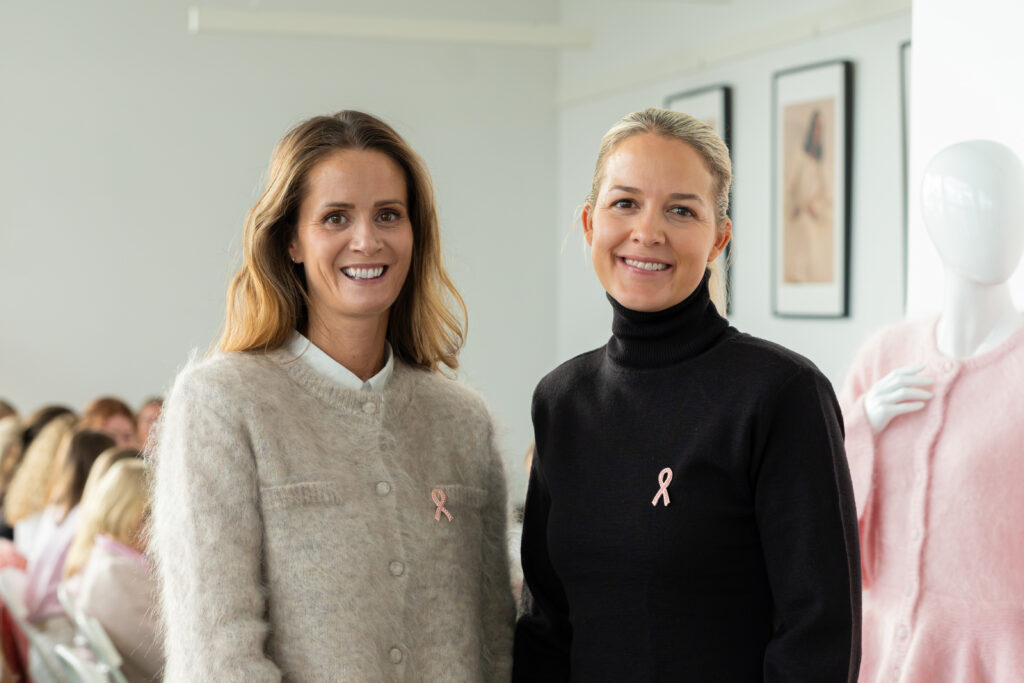 To kvinner kledd i gensere og med rosa sløyfe-brosjer står innendørs på et arrangement, med en utstillingsdukke og innrammede kunstverk i bakgrunnen.