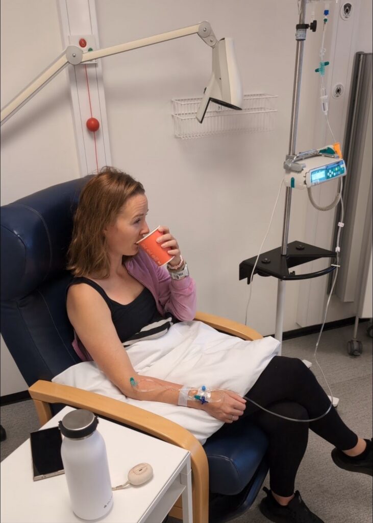 En kvinne sitter i en sykehusstol og får intravenøs behandling, mens hun nipper til noe fra en rød kopp. Ved siden av henne står medisinsk utstyr og et dryppstativ.