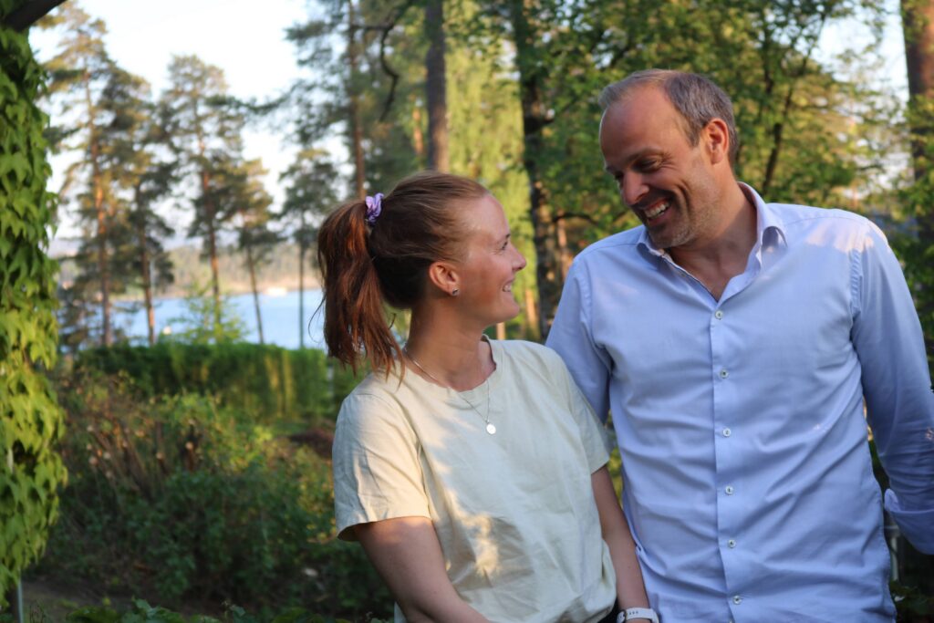 En kvinne og en mann står utendørs i et solfylt skogsområde og smiler til hverandre. I bakgrunnen ser man trær, grøntområder og et glimt av vann.