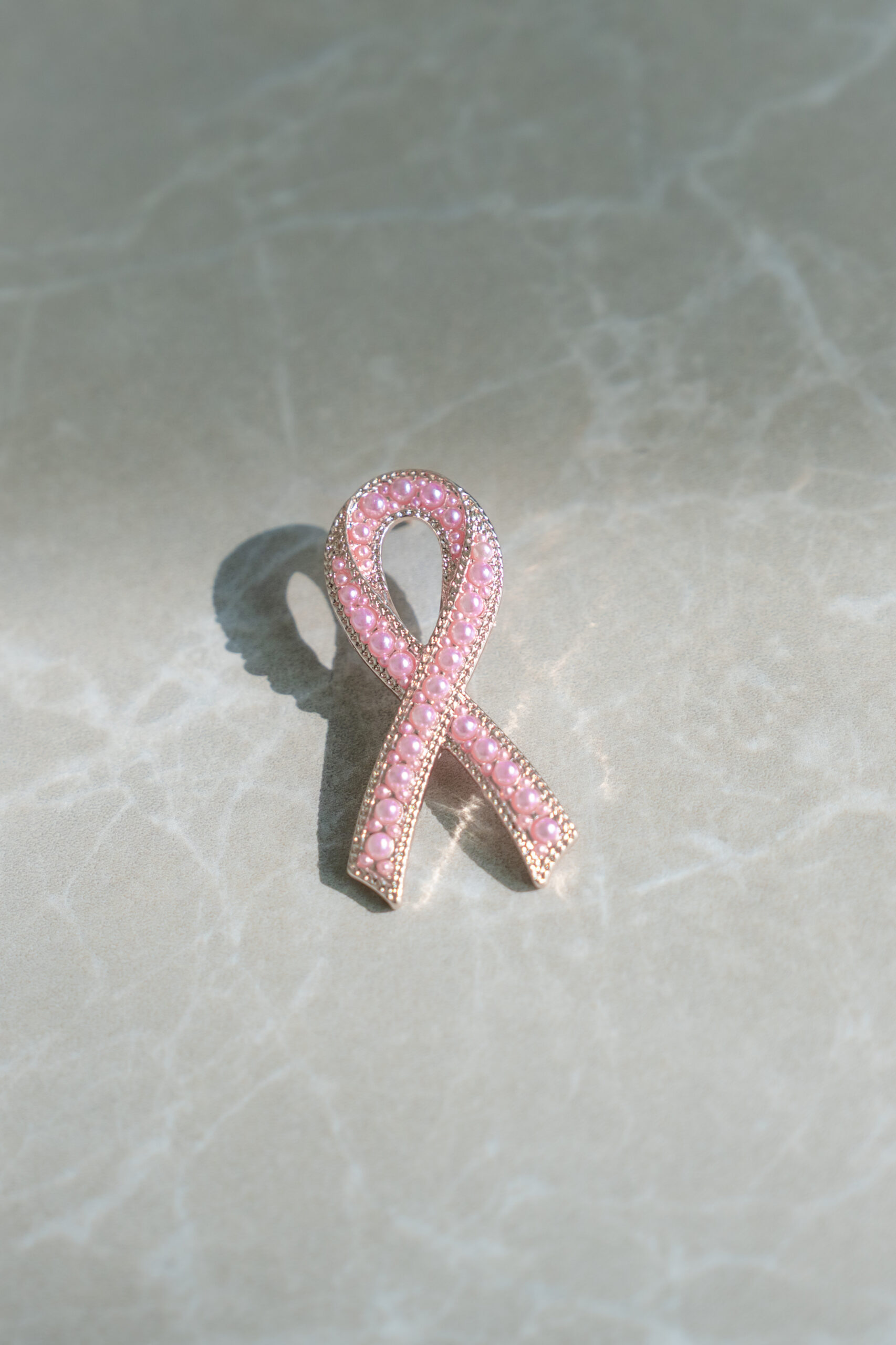 En brosje i form av et rosa bånd, prydet med små rosa edelstener, ligger på en lysegrå marmorflate.