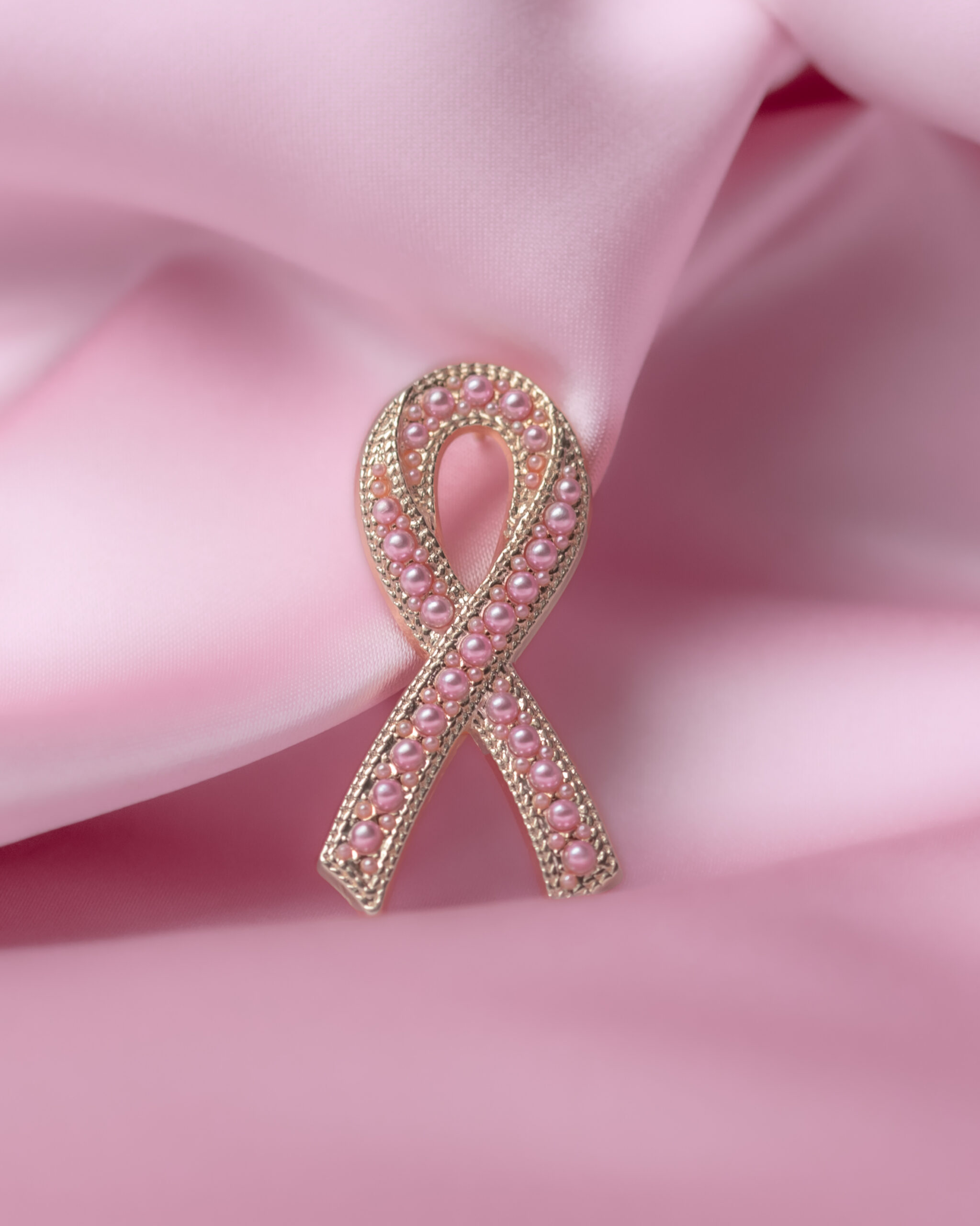 En rosa brosje med perler ligger på et mykt rosa stoff og symboliserer oppmerksomhet rundt brystkreft.
